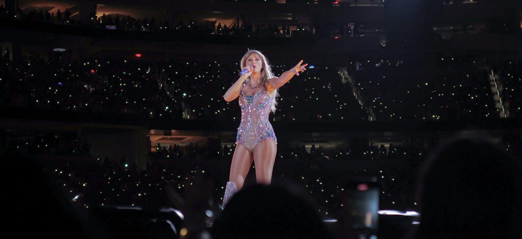Taylor Swift performs on stage during the Eras Tour, wearing a sparkling blue bodysuit adorned with sequins and matching knee-high boots, against a dark background with a spotlight illuminating her.