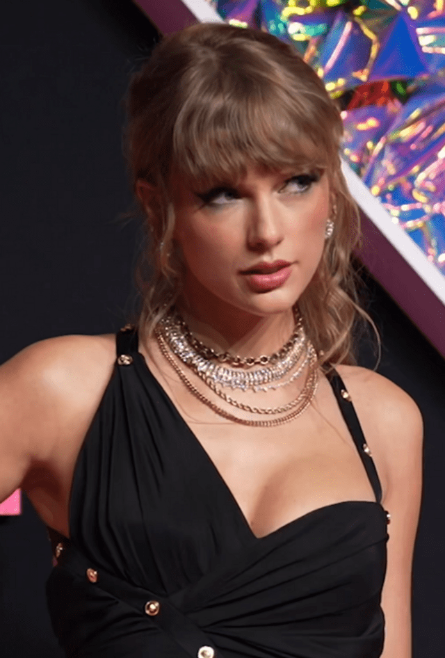 A close-up of Taylor Swift's face as seen at the MTV Video Music Awards. She is seen sporting a black dress, bangs, bold eye makeup, and five layers of thin gold and silver necklaces.