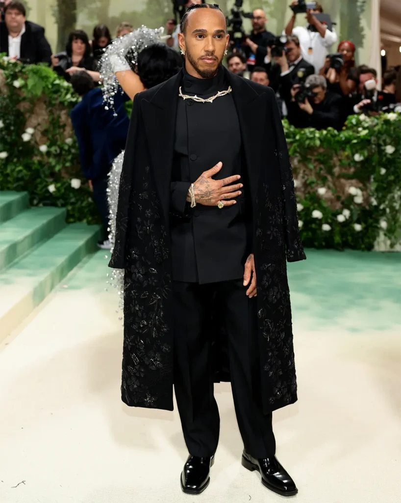 The image showcases Lewis Hamilton at the Met Gala 2024, making a fashion statement in an elegant black ensemble. He wears a tailored suit paired with an overcoat that features intricate floral embroidery, adding a touch of texture and sophistication to the outfit. His look is accessorized with a unique silver chain necklace that rests on the collar, enhancing the outfit's modern aesthetic. Lewis's tattoos are visible on his hands, which are adorned with stylish rings, adding a personal touch to his refined appearance. The background is filled with other gala attendees and numerous photographers, capturing the allure and grandeur of the event.