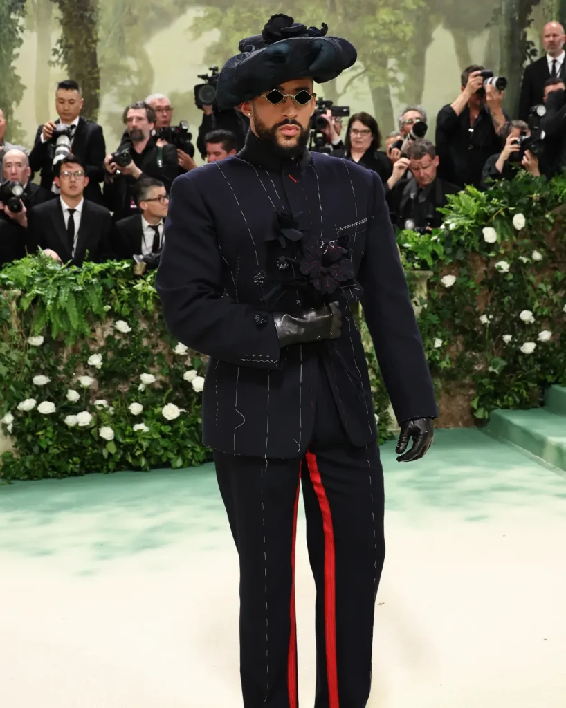 The image features  Bad Bunny at a Met Gala 2024, dressed in an avant-garde ensemble. His outfit consists of a dark, pinstriped suit with distinct stitching details and bold, red accents running down the sides of the trousers. The suit jacket is adorned with black floral motifs and he wears a large, sculpted hat in a matching style. His look is completed with black leather gloves and contemporary sunglasses. He carries a confident, serious expression, adding an intense aura to his striking fashion statement. In the background, numerous photographers capture his moment, underscoring the high-profile nature of the event.