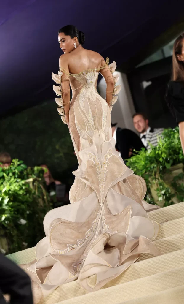 The image captures a Mona Patel  at the Met Gala 2024 , viewed from the back as she looks over her shoulder. She wears a striking gown with a creamy beige tone, intricately designed with floral embroidery and embellishments that shimmer under the event lighting. The dress features a dramatic, cascading train and structural, petal-like elements around the shoulders and down the back, creating a three-dimensional effect. Her hair is styled in a sleek updo, and she wears classic drop earrings. The backdrop is busy with other guests and photographers, highlighting the grandeur of the event.