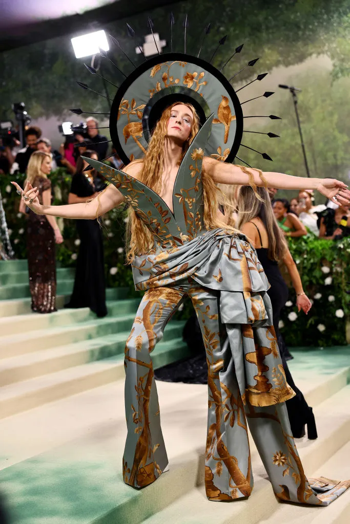 The image depicts a Harris Reed  at the Met Gala 2024 event, striking a dynamic pose in an artistically themed outfit. She wears a light gray ensemble with wide-leg trousers and a structured top, both adorned with golden botanical prints. The standout feature of her attire is a large circular halo-like headpiece with spokes, matching her outfit's color and theme, creating a sunburst effect. Her long blonde hair flows freely, and her arms are outstretched, adding to the dramatic flair of her presentation. The setting is bustling with onlookers and photographers capturing her moment, emphasizing the spectacle and grandeur of the occasion.