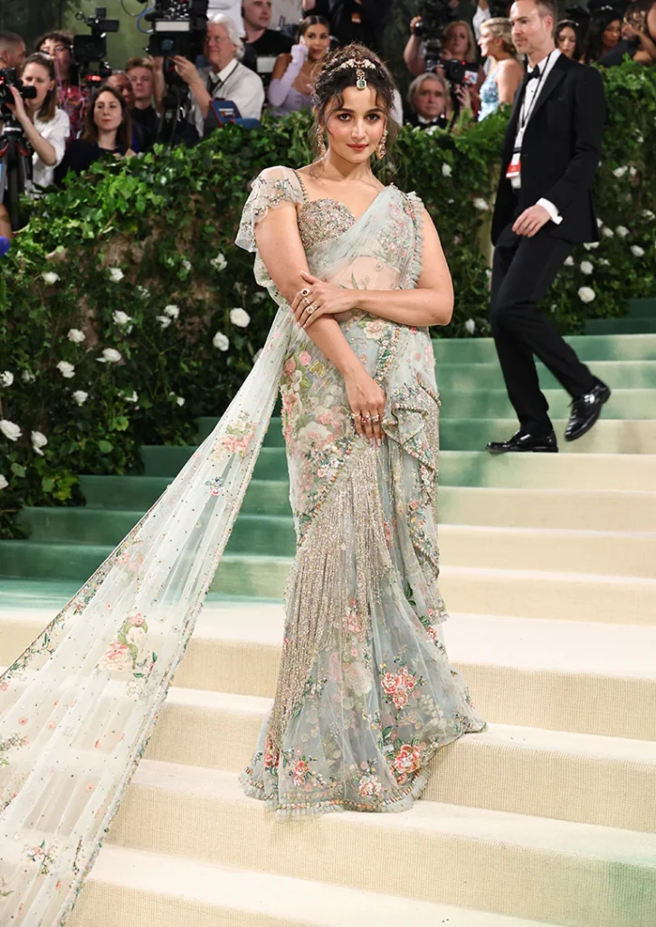 
The image captures Alia Bhatt at the Met Gala 2024, elegantly clad in a traditional-inspired gown that gracefully combines modern fashion with cultural heritage. Her outfit features a pastel floral sari embellished with intricate beadwork and sequins, adding a radiant glow to her appearance. The sari is paired with a long, flowing train that adds a dramatic touch to her ensemble. Her look is complemented with a delicately jeweled headpiece, enhancing her regal bearing. Alia's makeup is soft and enhances her natural beauty, while her hairstyle is swept back, allowing the details of her outfit to shine. Surrounded by onlookers and photographers, she exudes confidence and grace on this grand occasion.