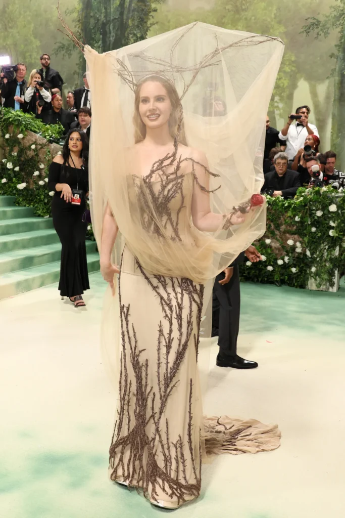 The image features a Lana Del Ray at a glamorous Met Gala 2024 event, dressed in a unique gown with a nature-inspired theme. She wears a delicate sheer veil that flows over her head and shoulders, held aloft by a crown of realistic branches. The dress itself is adorned with dark, branch-like embroidery that cascades down the front and along the gown's length, mimicking roots and natural growth. Her makeup is natural with a hint of sophistication, complementing her radiant smile. In the background, photographers and other attendees capture the moment, adding to the lively atmosphere of the event.