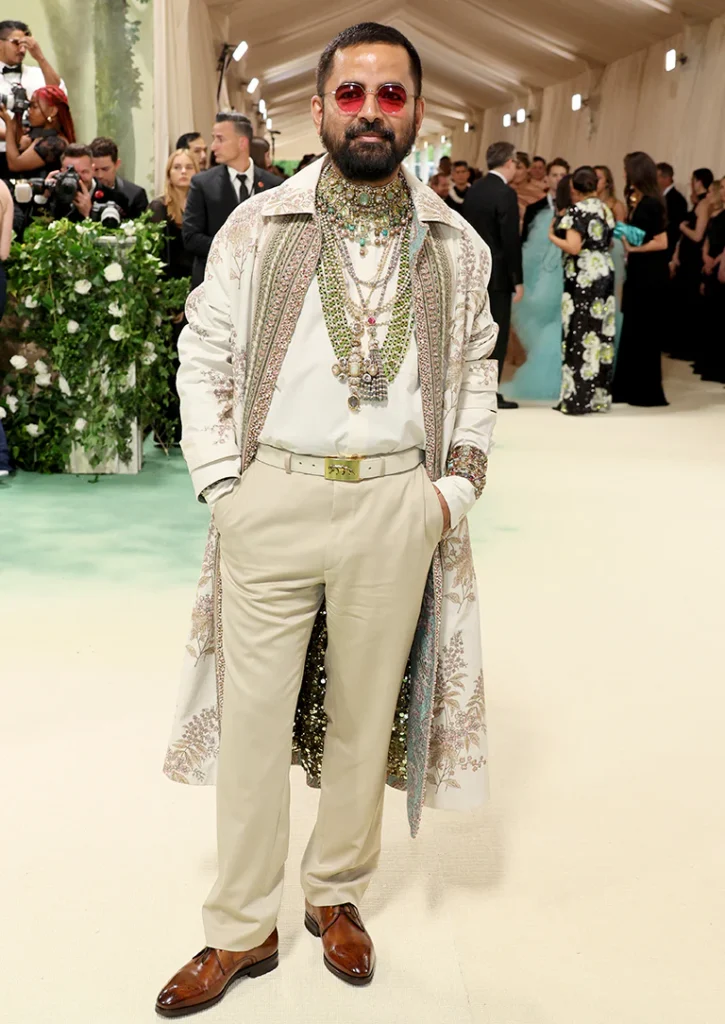 The image showcases Sabyasachi Mukherjee at the Met Gala 2024, embodying a sophisticated and ornate style. He is dressed in an exquisite cream-colored ensemble featuring a detailed, embroidered jacket with embellished accents that capture the light, creating a luxurious appearance. He wears multiple layers of elaborate jewelry, including necklaces with precious stones and intricate designs that highlight his cultural heritage. His look is completed with red-tinted sunglasses, adding a contemporary edge to his traditional attire. Sabyasachi stands confidently, surrounded by other attendees, which emphasizes the vibrancy and grandeur of the event.
