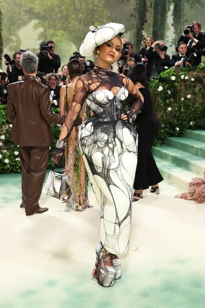 The image captures Natasha Poonawalla at the Met Gala 2024, donned in a bold and artistic outfit. Her ensemble features a form-fitting bodysuit with a sheer overlay, adorned with abstract, monochromatic designs that create an eye-catching visual illusion. The designs resemble ink blots or paint smears, lending an avant-garde and surreal feel to her look. Complementing her attire is an extravagant white hat, sculpted to mimic cloud formations or foam, adding to the theatrical impact of her outfit. Natasha’s makeup is dramatic, with bold eyes and neutral lips, which harmonize with her striking ensemble. She poses confidently against the backdrop of photographers and gala attendees, showcasing her unique fashion statement.