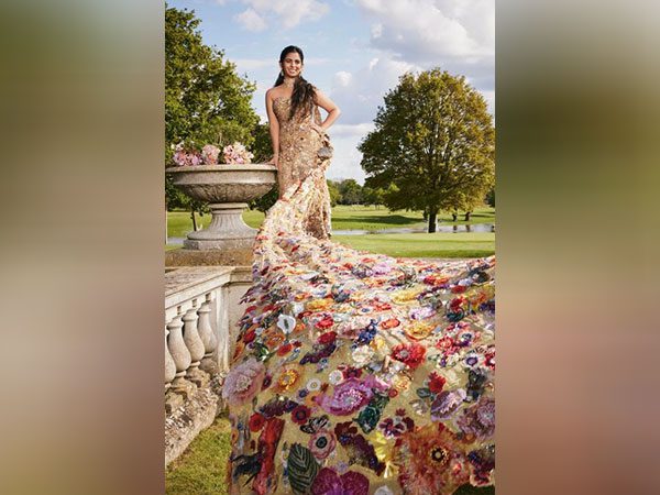 The image showcases Isha Ambani at the Met Gala 2024, wearing a stunningly intricate gown that is a true work of art. The top part of the dress fits snugly and is adorned with detailed golden embroidery that enhances her elegant silhouette. This transitions into a lavish, full-length skirt decorated with a vibrant mosaic of floral appliqués, each meticulously crafted with a mix of rich colors and textures. The dress's long train spreads across the grass, providing a breathtaking visual that blends harmoniously with the lush outdoor setting. Isha stands near a classical stone urn overflowing with flowers, complementing her gown's floral theme. Her hair is styled in a sleek, simple manner, allowing the dress to take center stage, and her poised demeanor reflects the grandeur of the occasion.