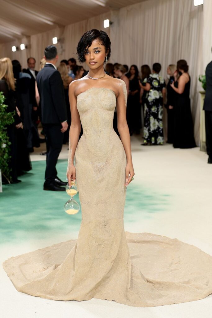 The image shows Tyla standing poised at the Met Gala event, wearing a sculpted gown that mimics the appearance of sand. The dress is strapless, fitted through the bodice, and flares into a flowing train that puddles elegantly at her feet, textured to resemble a sandy shore. She holds an hourglass in one hand, which complements the theme of her attire, emphasizing the passage of time. Her makeup is subtle yet sophisticated, with a focus on her eyes, and her hair is styled in a short, wavy bob. The setting includes other guests in the background, highlighting her standout presence on a busy and vibrant occasion.