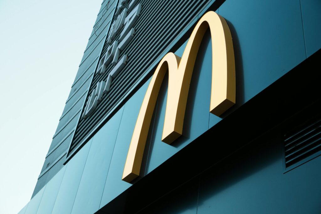 A low-angle shot of the McDonald’s logo, the iconic golden arches, against the backdrop of a modern building exterior. The sky is clear, and the structure emphasizes the large, bright logo.