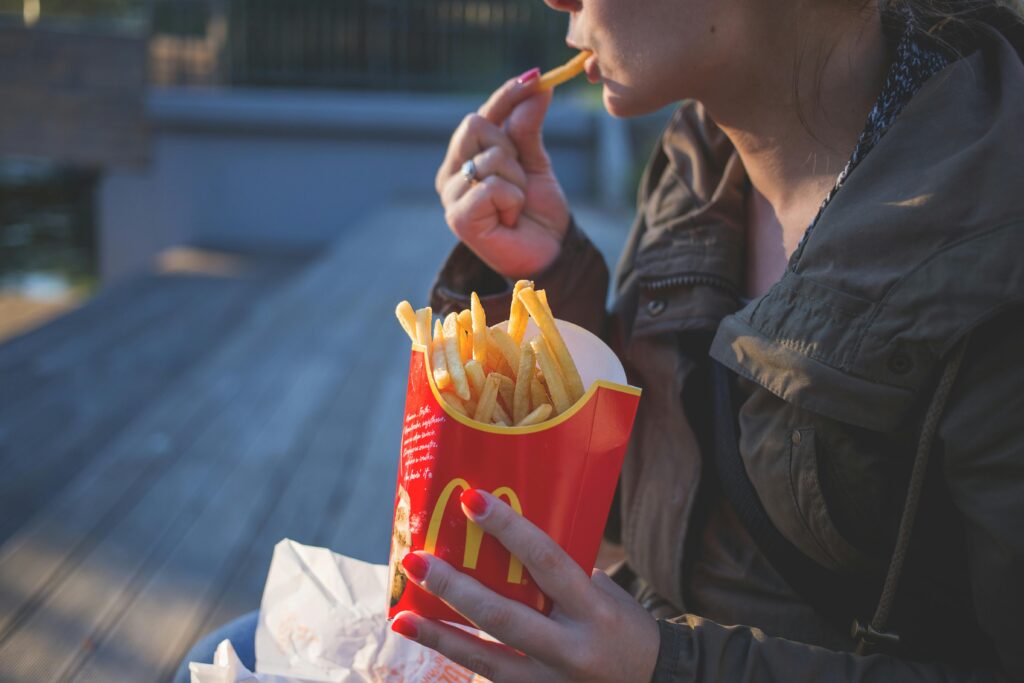 A close-up of a person eating McDonald’s French fries outdoors, holding a red McDonald’s fries box. The person is dressed in a jacket, and the scene appears to be relaxed with natural light illuminating the setting.