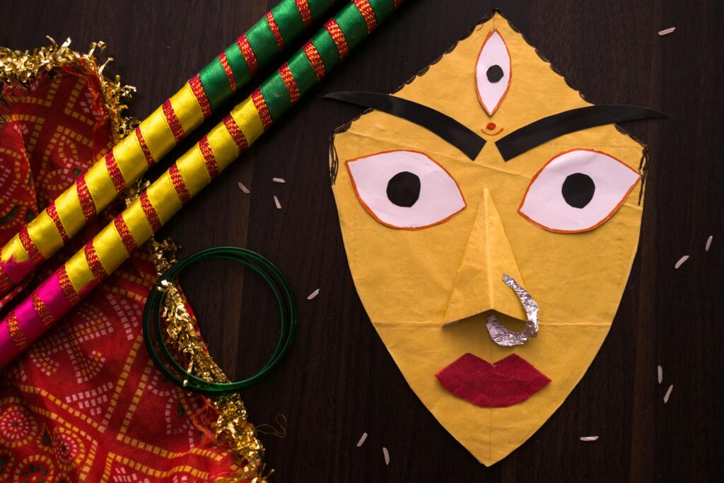 A handmade paper mask of Goddess Durga placed on a table alongside traditional Indian textiles, bangles, and decorative sticks used for Durga Puja celebrations.