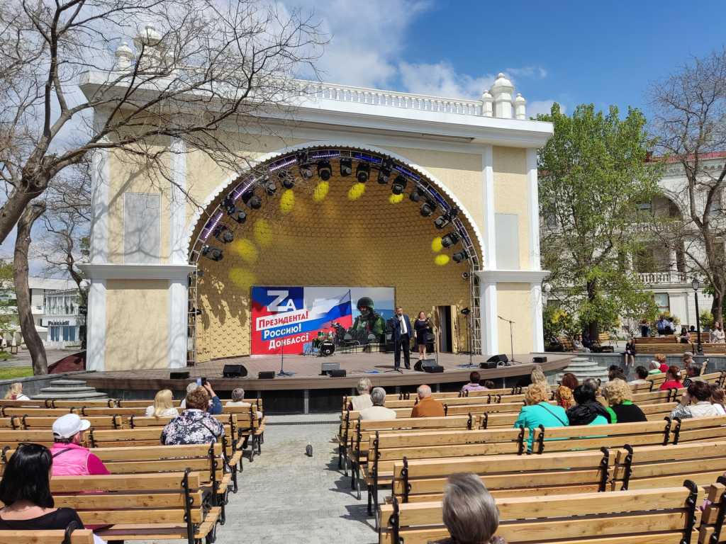 An outdoor propaganda rally in Sevastopol, April 2022, with a stage featuring banners supporting the Russian invasion of Ukraine, including the slogan ‘For the President! For Russia! For Donbas!’ under a clear sky.