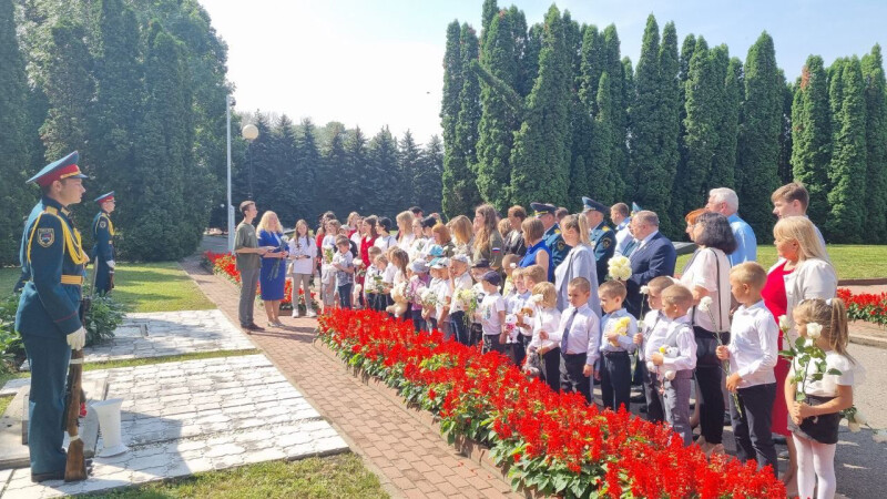 A crowd of children and adults holding flowers during a memorial ceremony, guarded by soldiers in uniform, against a backdrop of lush greenery and red flower arrangements, commemorating those lost in conflict.