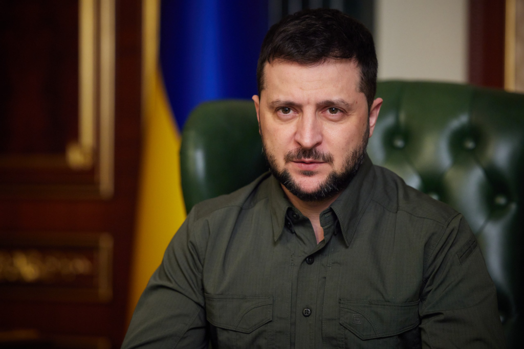 Ukrainian President Volodymyr Zelensky seated in a green leather chair with the Ukrainian flag in the background, wearing a green military-style shirt, conveying a solemn and determined expression